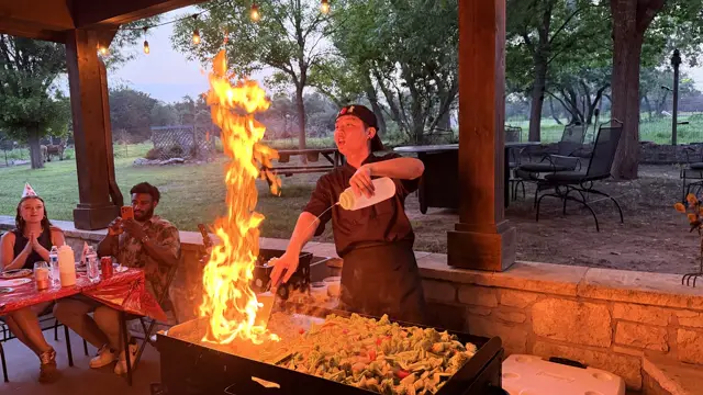 A tower of flame rises off the grill as the chef pours oil at a dusk party
