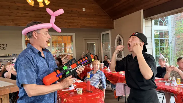 A guest in a balloon hat sprays the chef with a water gun